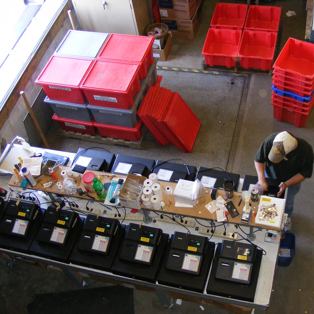 A worker standing at a long table, assembling electronic devices in a warehouse with stacks of red bins nearby.