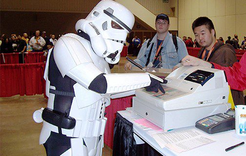 A person dressed as a stormtrooper from star wars uses a photocopier at a convention, with two men observing.