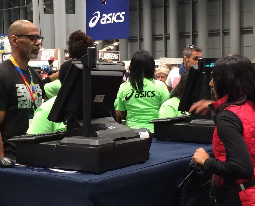 People at a registration desk, wearing bright green shirts marked "volunteers," inside a busy event hall with an asics logo visible.