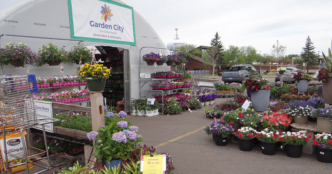 Outdoor garden center with colorful flowers and plants displayed under a white tent labeled "garden city," with a selection of potted plants and price tags visible.