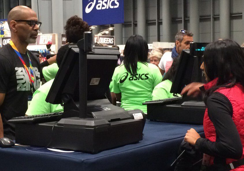 People at a registration desk, wearing bright green shirts marked "volunteers," inside a busy event hall with an asics logo visible.